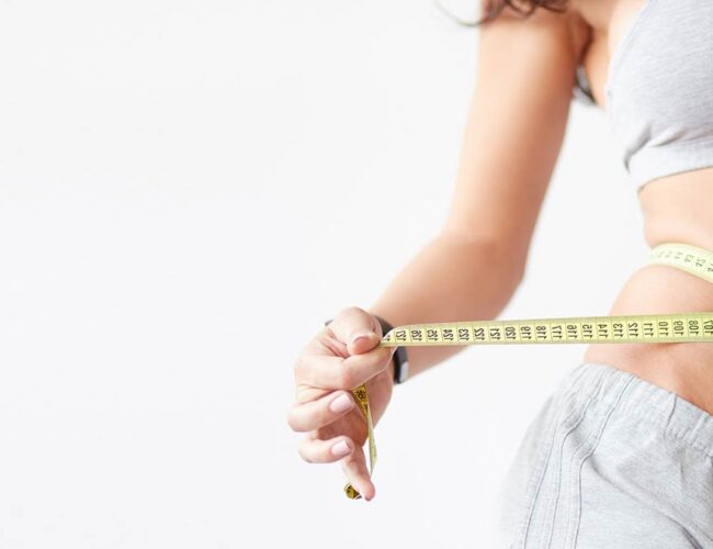 Woman in athletic wear measuring her waist with a yellow tape measure, symbolizing weight loss and digestive health benefits of natural colon cleanse.