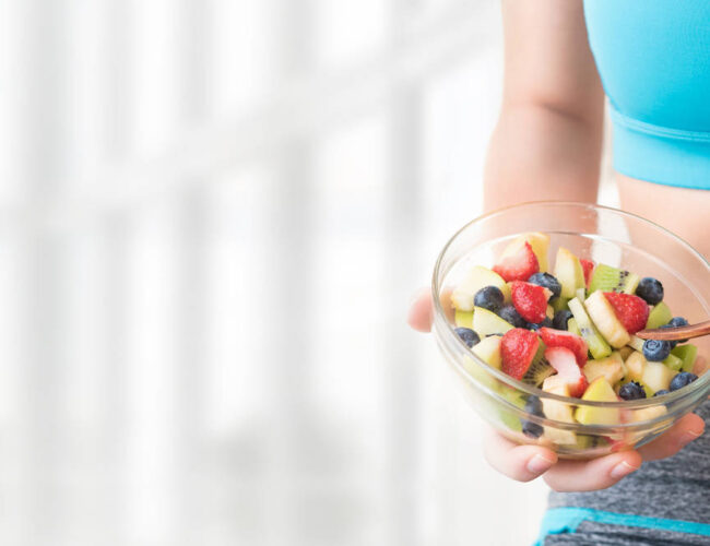 Woman in fitness wear holding a bowl of fresh fruit salad, symbolizing healthy digestion and colon hydrotherapy benefits at Amber Colonics.