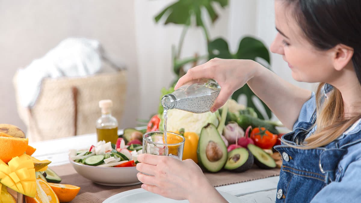 Woman pours water into a glass beside a table of fresh fruits and vegetables, supporting hydration and gut comfort.