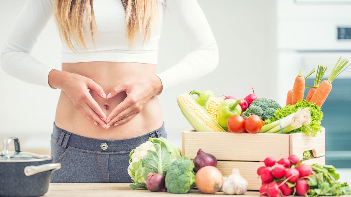 Woman holding hands over stomach next to fresh vegetables and healthy foods, representing colon hydrotherapy preparation and digestive wellness in Tempe
