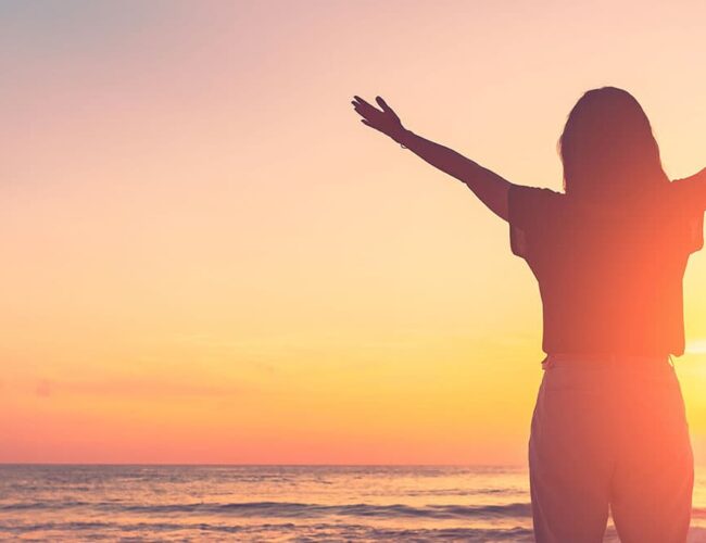 Woman welcoming the sunrise by the water, representing a renewed fall wellness routine