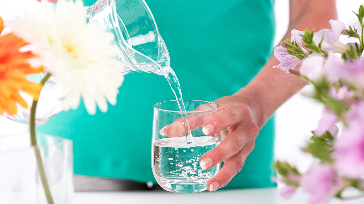 Person pouring water from a glass pitcher into a drinking glass beside flowers.